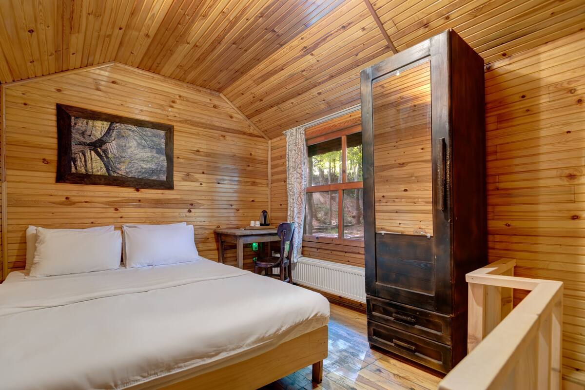 A wooden cabin bedroom with a bed, a desk, and a large window.