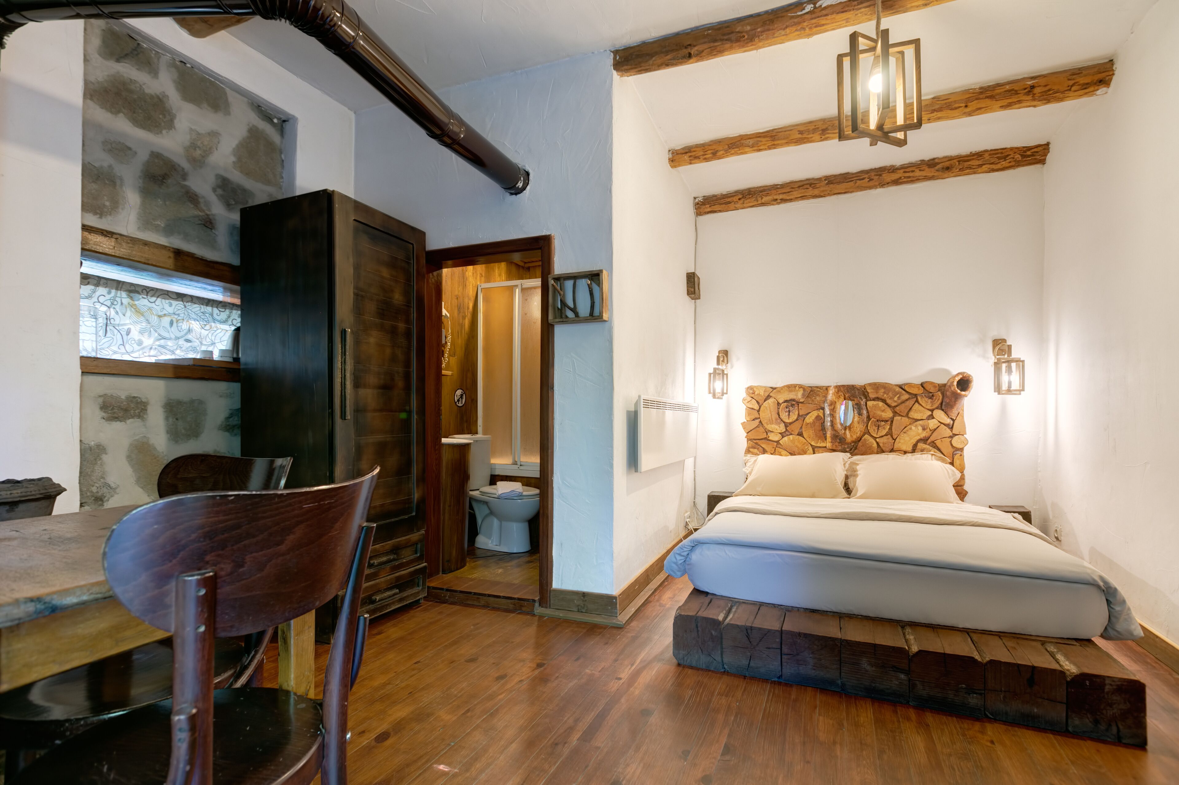 Ein Schlafzimmer mit Stein-Kopfstütze, Holzbalken, einem Bett, einem Holztisch mit Stühlen und einem Fenster zur Steinwand.