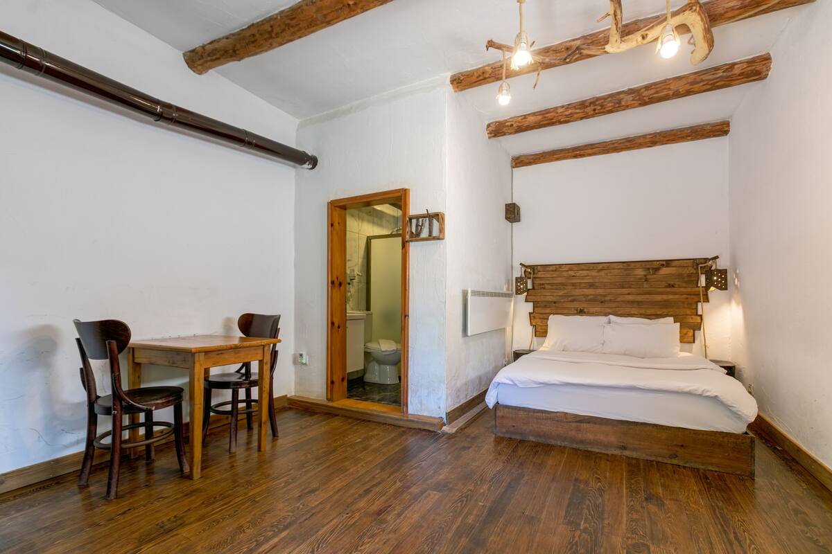 A bedroom with a wooden bed, a wooden table with chairs, and a bathroom visible through an open door.