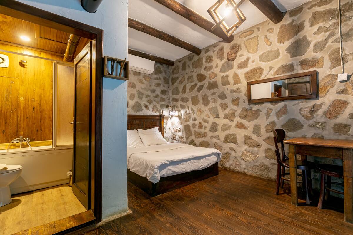 A bedroom with a wooden bed, stone walls, a wooden table, and a mirror.