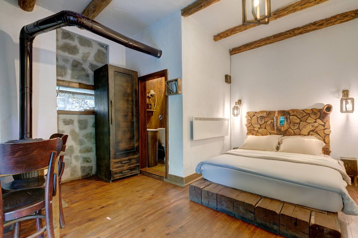A bedroom with a stone headboard, wooden beams, a bed with white linens, a wooden chair, and a stone wall.
