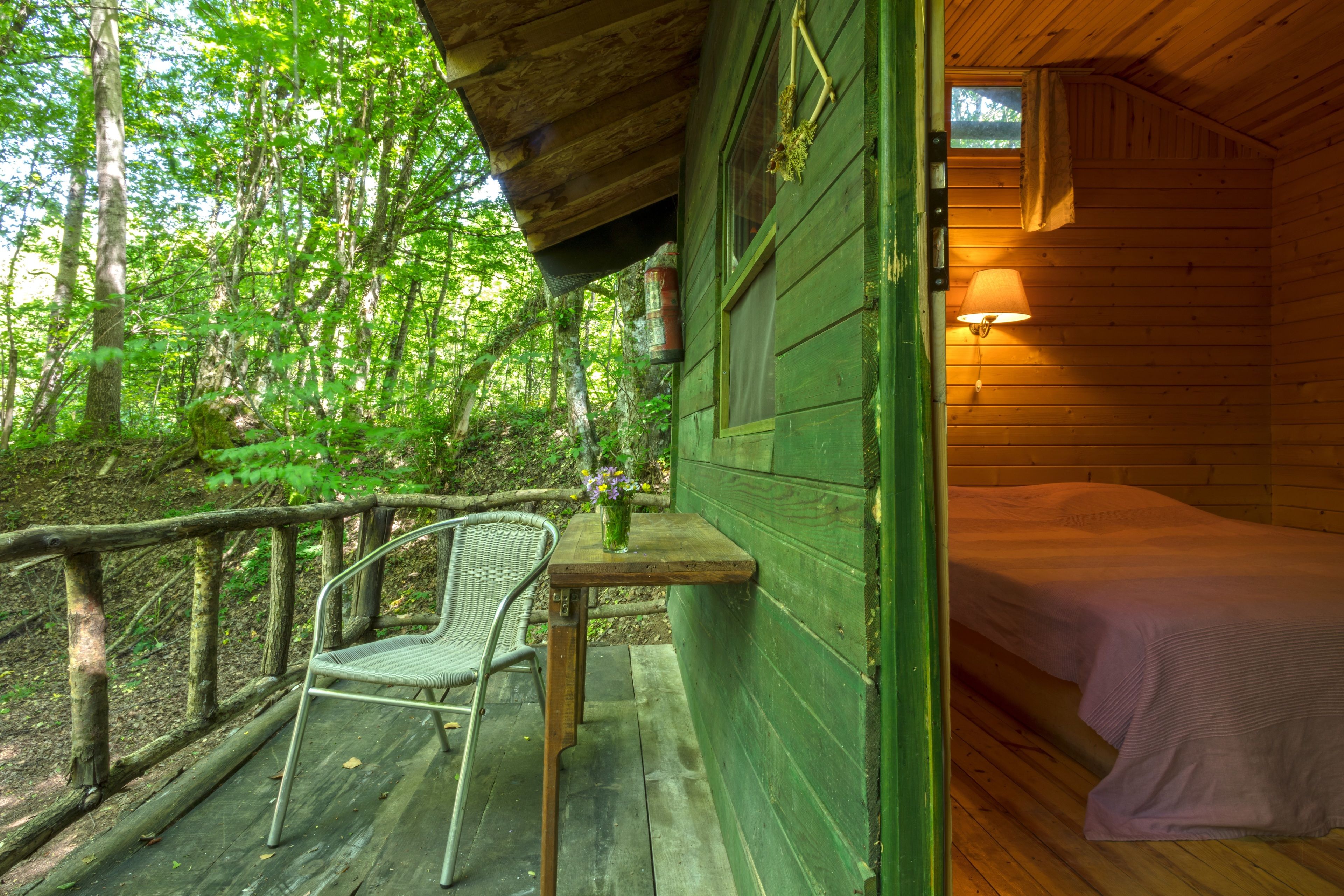 Uma cabana de madeira com uma cama, uma mesa e uma cadeira num deck rodeado por árvores.
