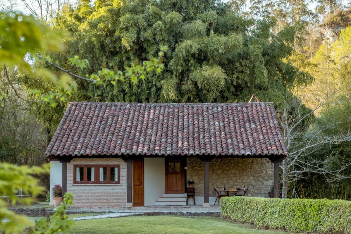 A rustic house with a tiled roof, a stone wall, and a small garden with a pond.