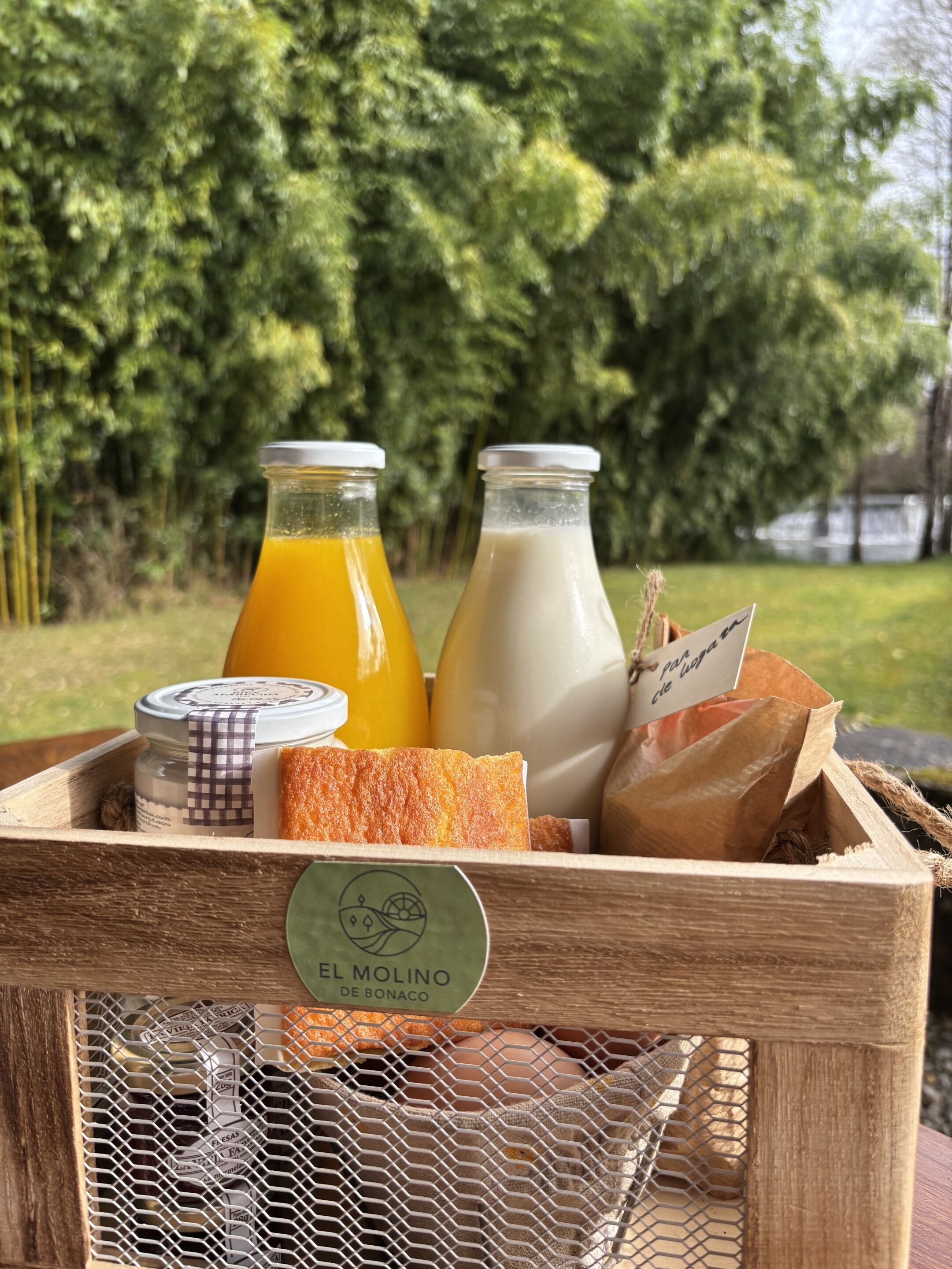 Una caja de madera con frente de malla que exhibe la etiqueta 'EL MOLINO DE BONACO', contiene dos botellas de vidrio con jugo de naranja y leche, un pan y una bolsa de papel con una etiqueta.
