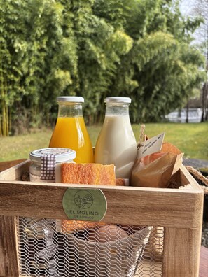A wooden crate with a mesh front labeled "EL MOLINO DE BONACO," containing two glass bottles of orange juice and milk, a loaf of bread, and a paper bag with a tag.