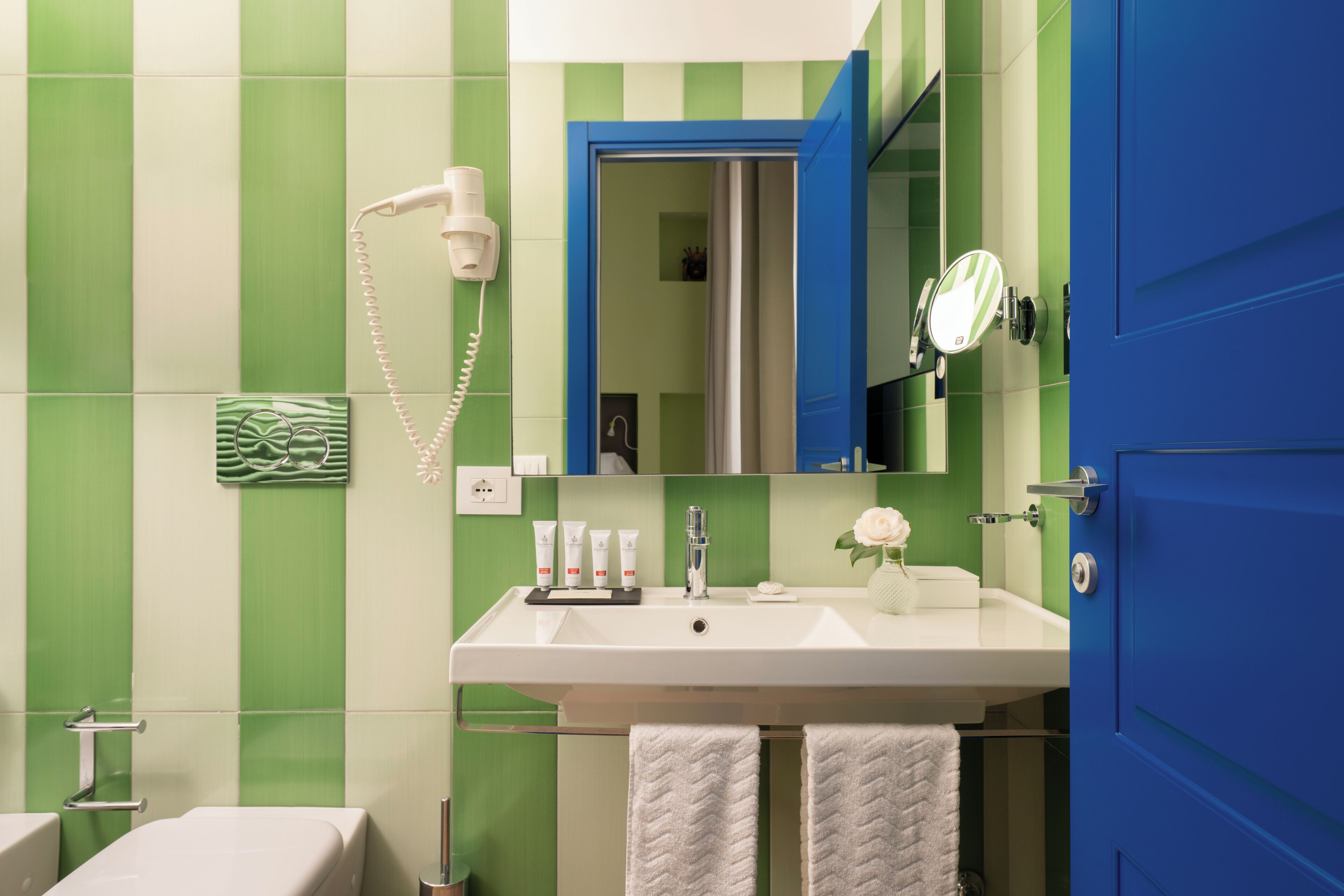 Een badkamer met een witte wastafel, muren met groene en witte strepen, een blauwe deur, een haardroger, een spiegel en twee handdoeken.