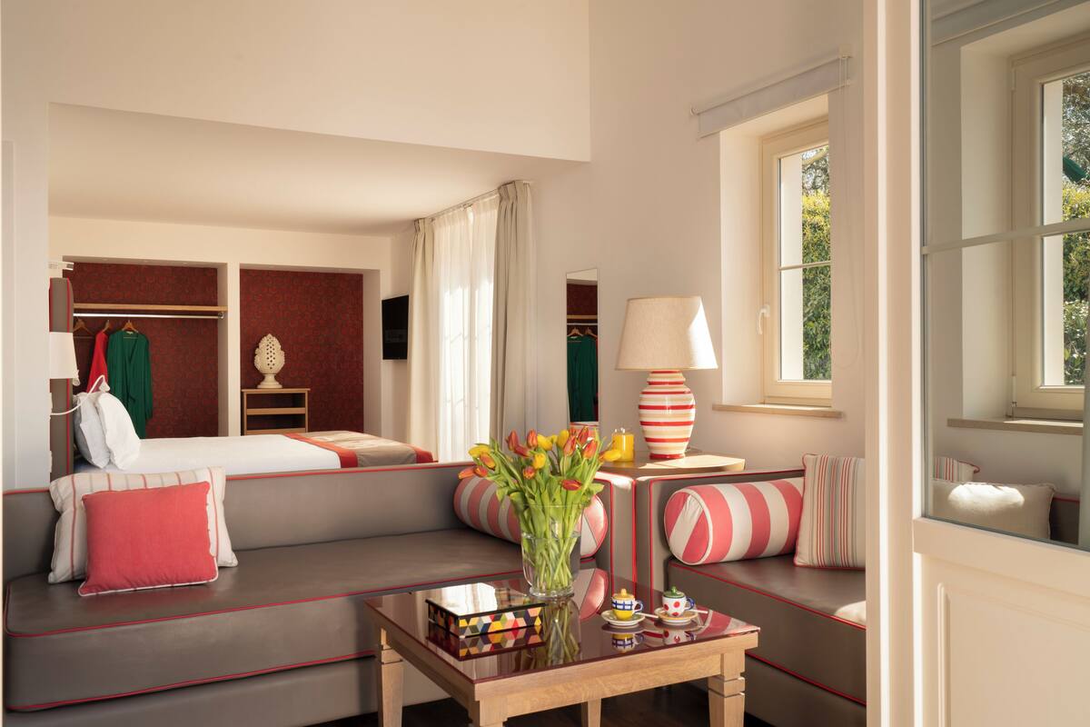 Une chambre d’hôtel moderne, dotée d’un canapé, d’une petite table ornée d’un vase de fleurs, d’une lampe et offrant une vue sur la verdure environnante.