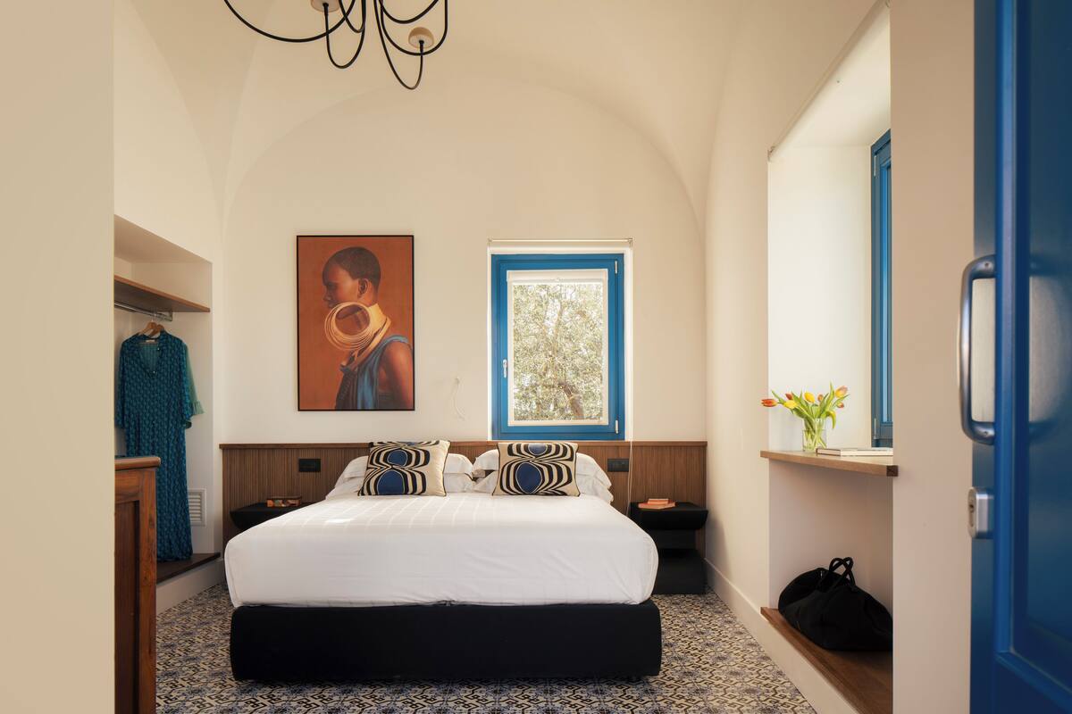 Une chambre avec un grand lit, une fenêtre donnant sur des arbres, un tableau au mur et une porte bleue.