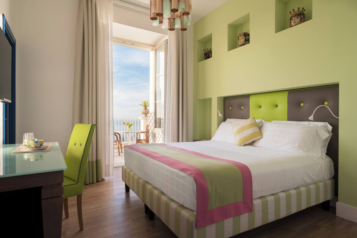 Une chambre d’hôtel avec un lit, un bureau et une chaise. Elle dispose d’un balcon avec vue sur le ciel.