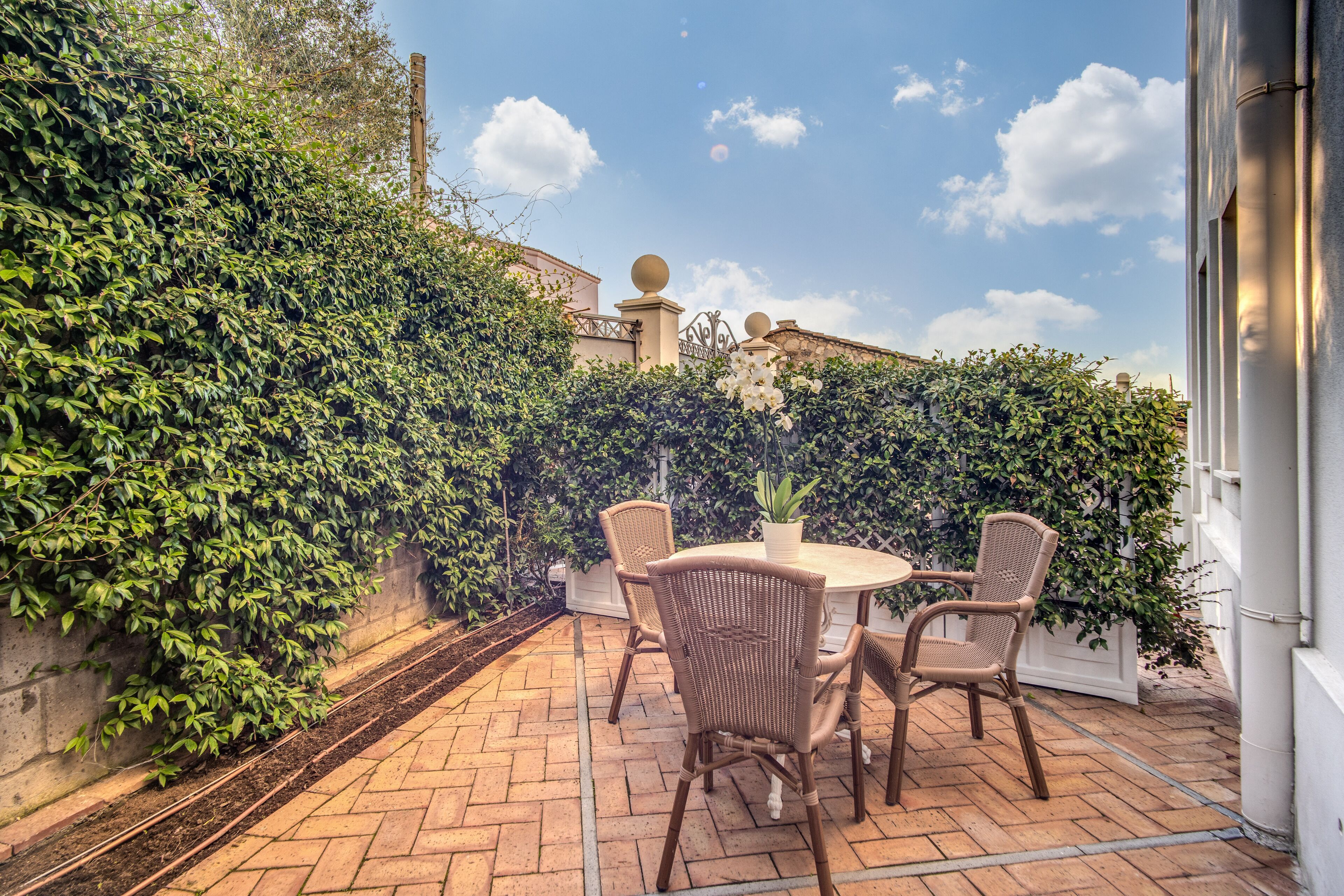 An outdoor patio with a round table and wicker chairs, surrounded by lush greenery and a brick floor.