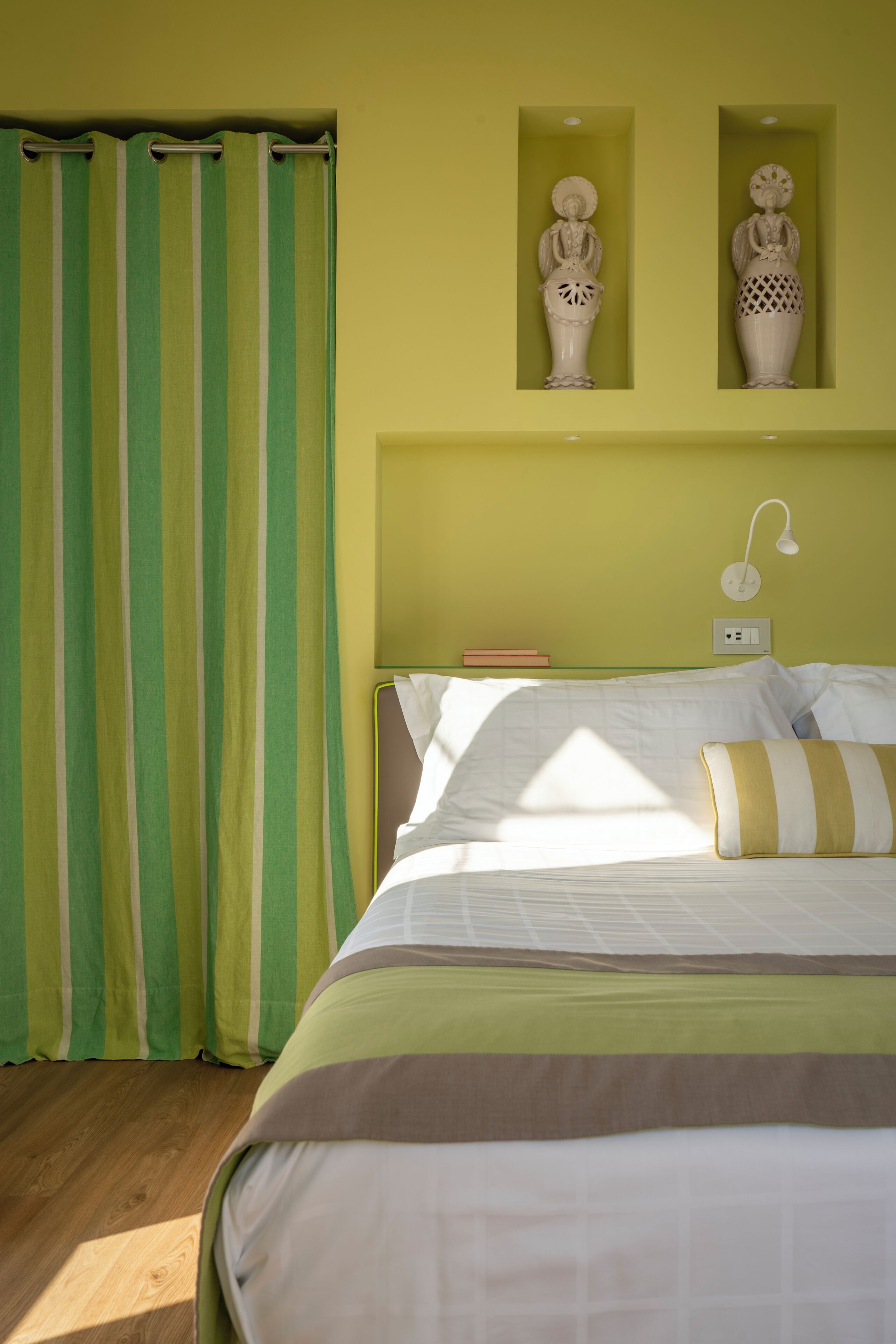 Une chambre avec un lit, un rideau à rayures vertes et blanches, deux vases accrochés au mur et une lampe de chevet.