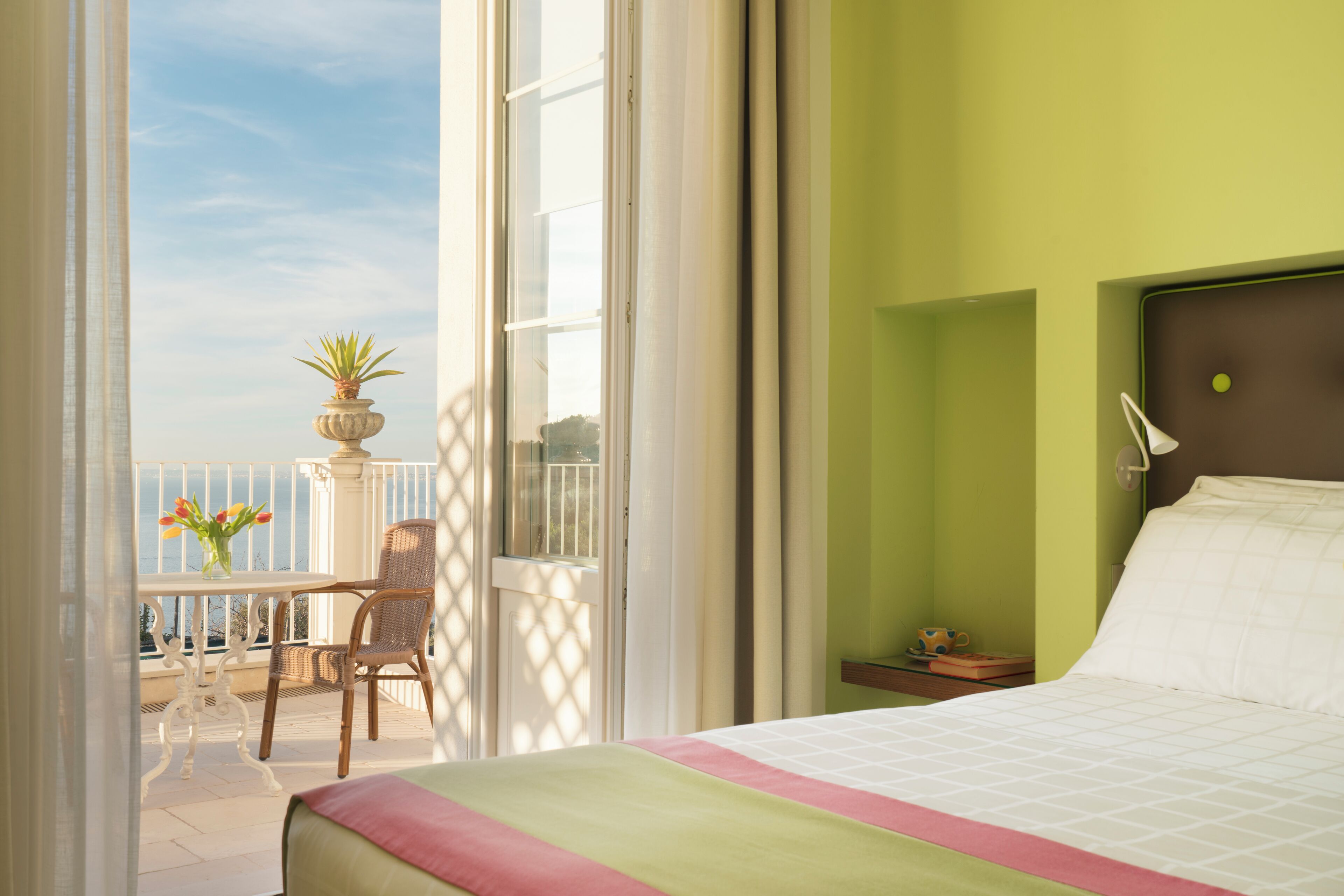 A hotel room with a balcony, a bed with a green and pink bedspread, a wicker chair, and a small table with a vase of flowers.