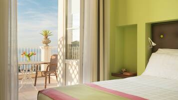 A hotel room with a balcony, a bed with a green and pink bedspread, a wicker chair, and a small table with a vase of flowers.