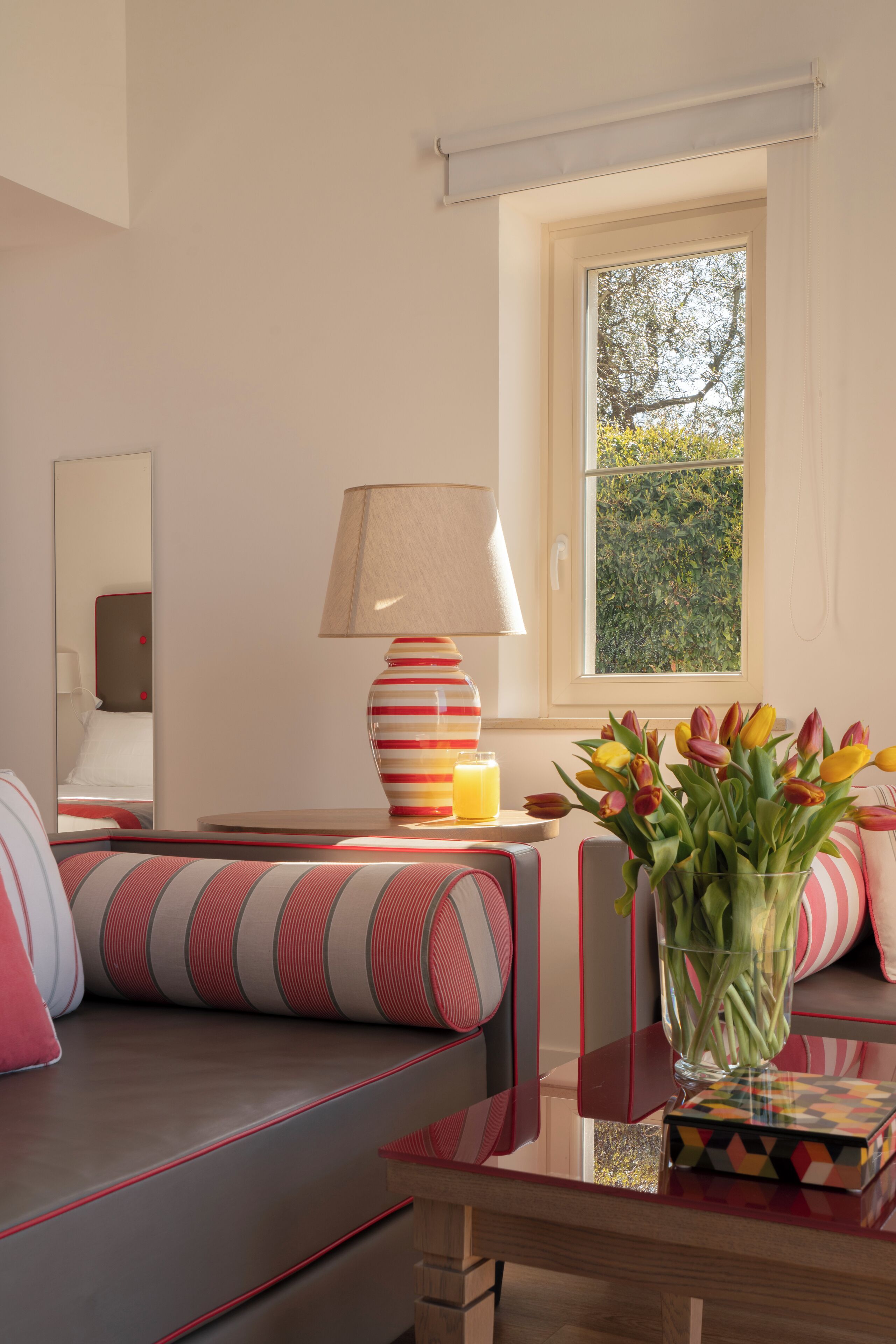 A modern hotel room with a sofa, a lamp, a vase with flowers, and a window with a view of greenery.