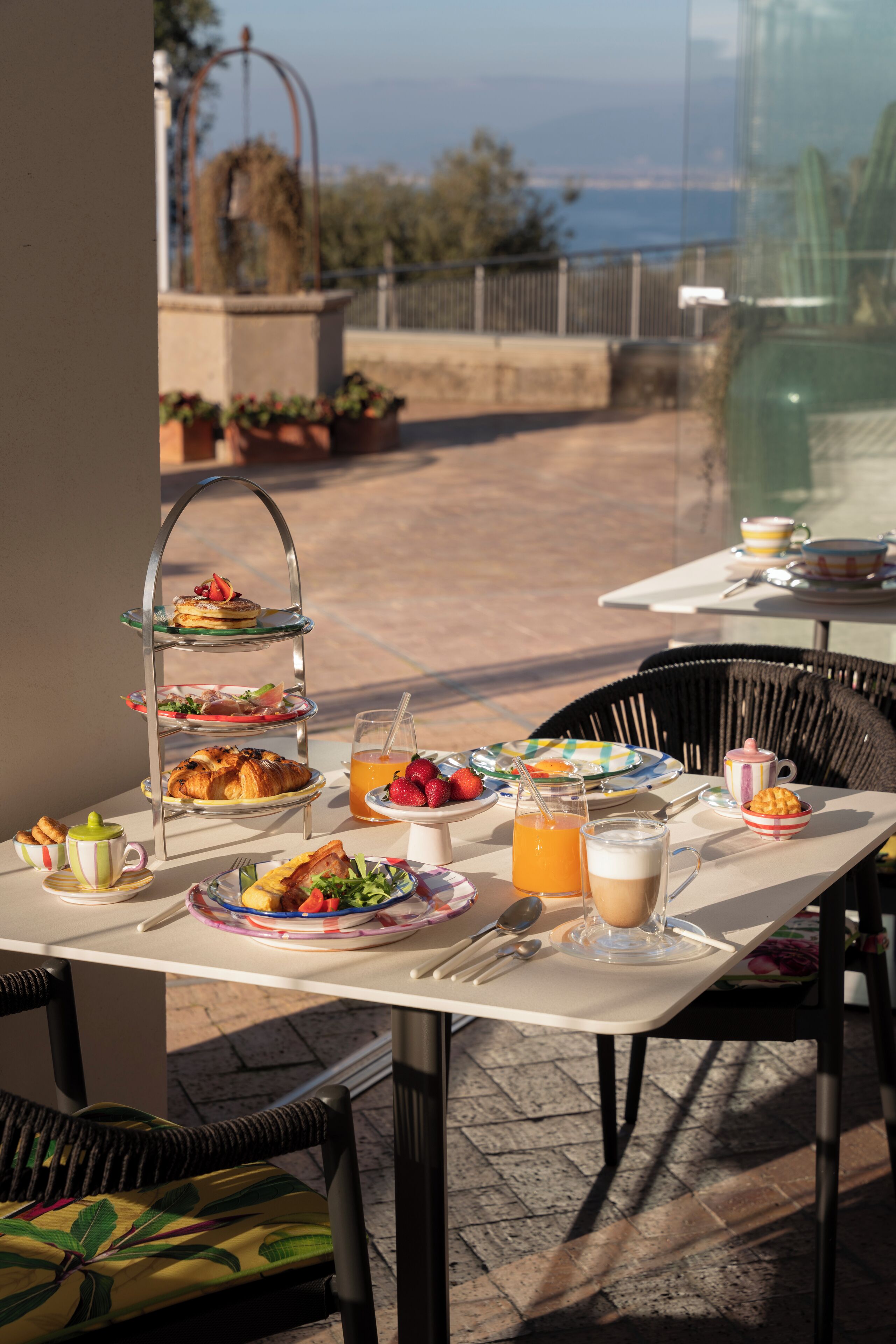 A three-tiered dessert stand with pastries, a table with breakfast items including eggs, toast, and fruit, and a view of the sea through a glass window.
