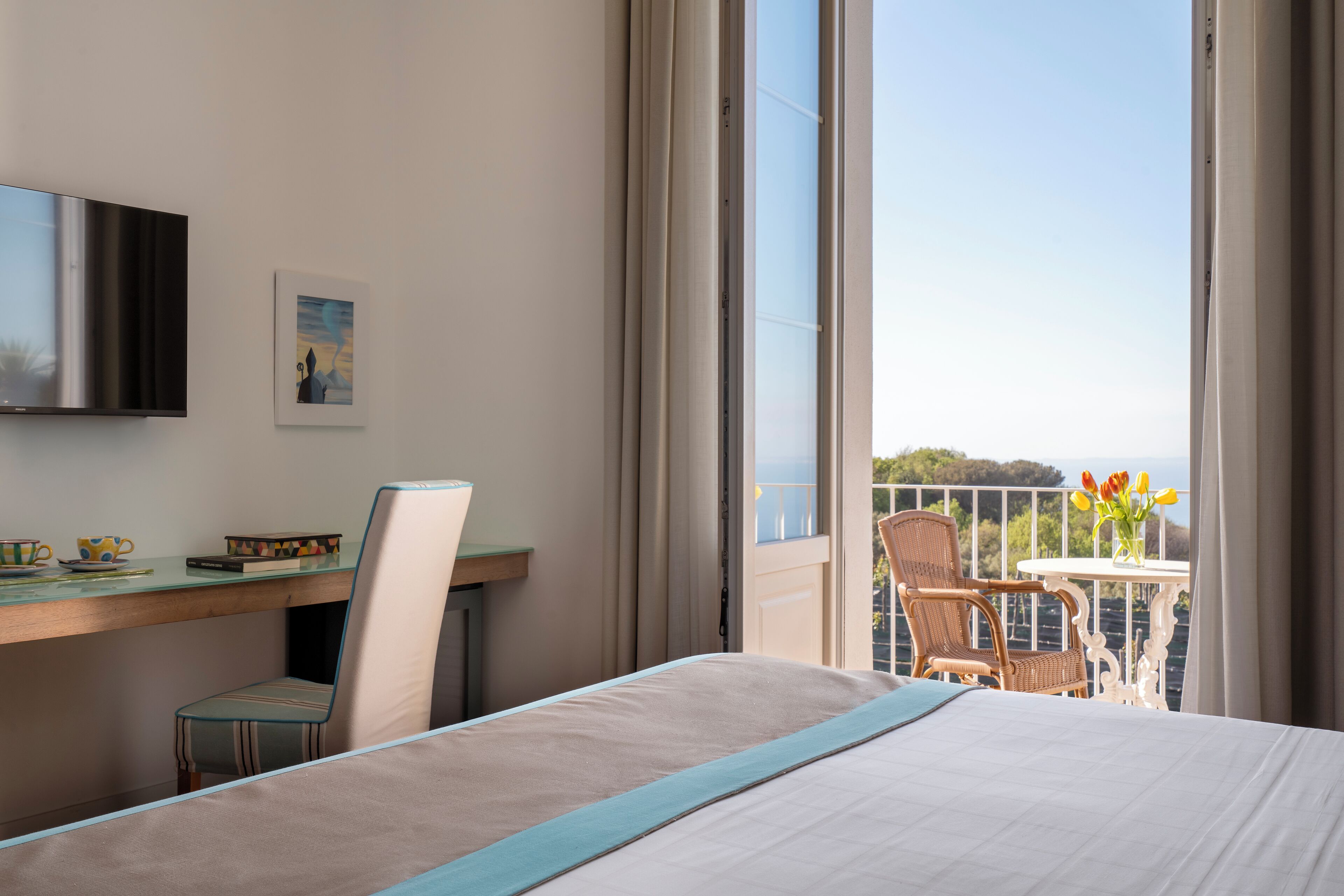 Et hotelværelse med en seng, et skrivebord med stol, et fjernsyn og en balkon med bord og stole.