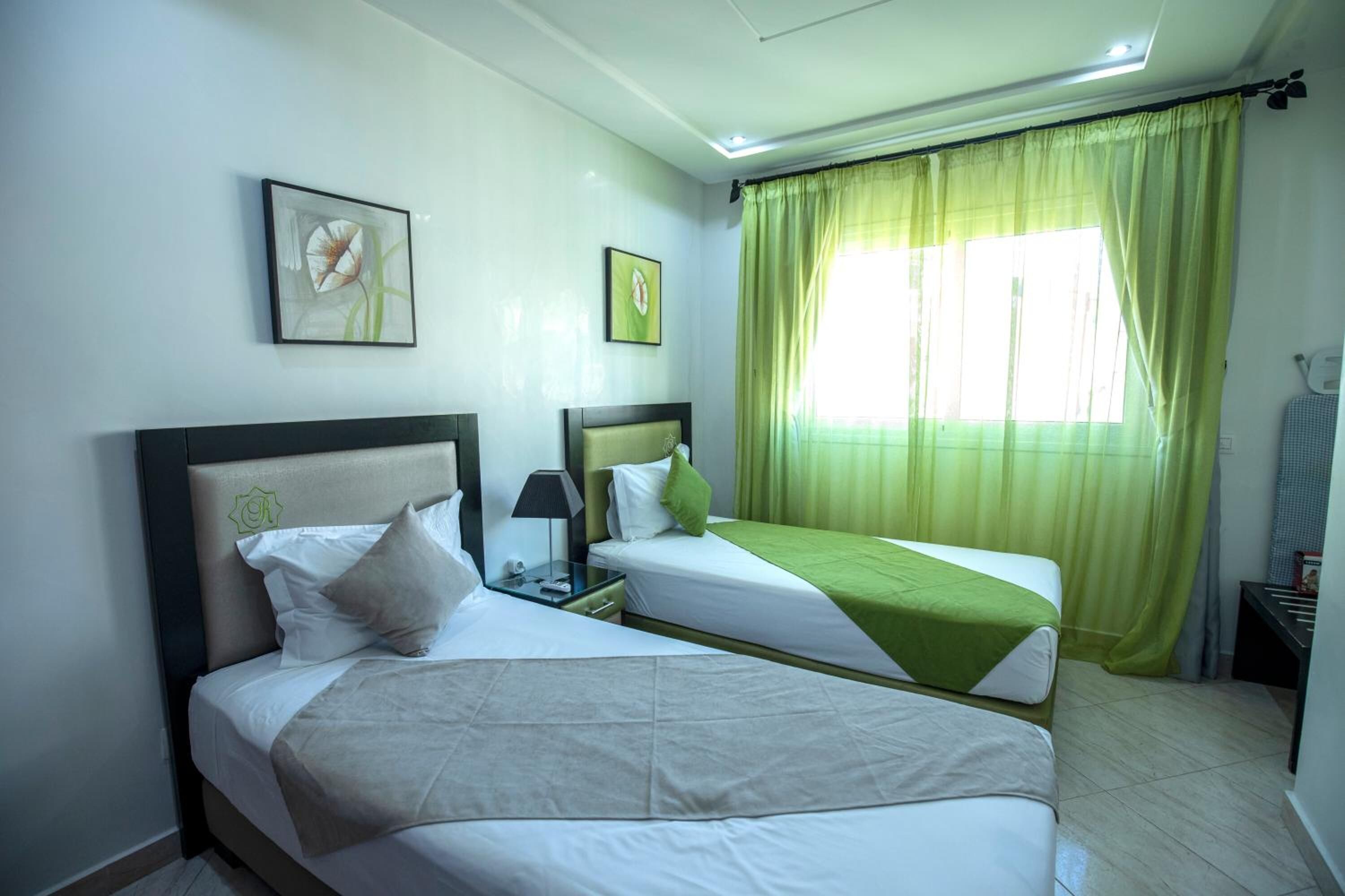 A hotel room with two beds, a nightstand, a lamp, and a window with green curtains.