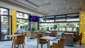 Un área de estar interior moderna con mesas, sillas, ventanas amplias y una pantalla de televisión.