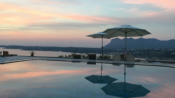 An infinity pool with two umbrellas, chairs, and a view of a body of water and mountains. - Theodosia Studios (Chania)