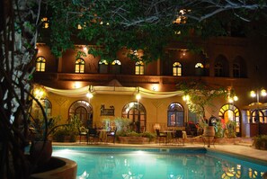 Zona junto a la piscina con comedor y un edificio de ventanas arqueadas e iluminación cálida.