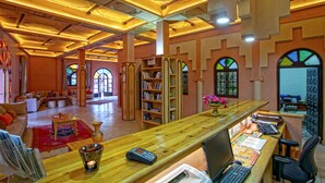 A spacious lobby with a wooden reception desk, seating area, and bookshelves. - Kasbah Sirocco (Zagora)