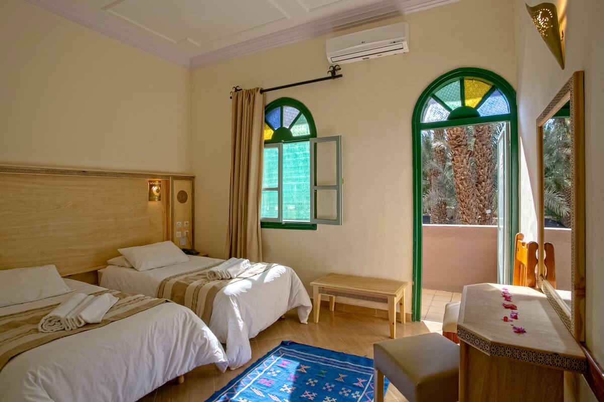Habitación de hotel con dos camas, un banco, una mesita con espejo y un ventanal con marco verde.