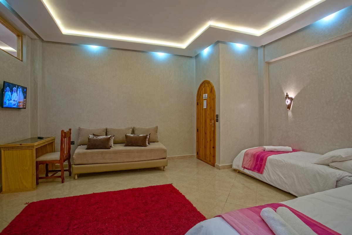 Habitación de hotel con sofá, dos camas, alfombra roja, escritorio de madera y televisor en la pared.