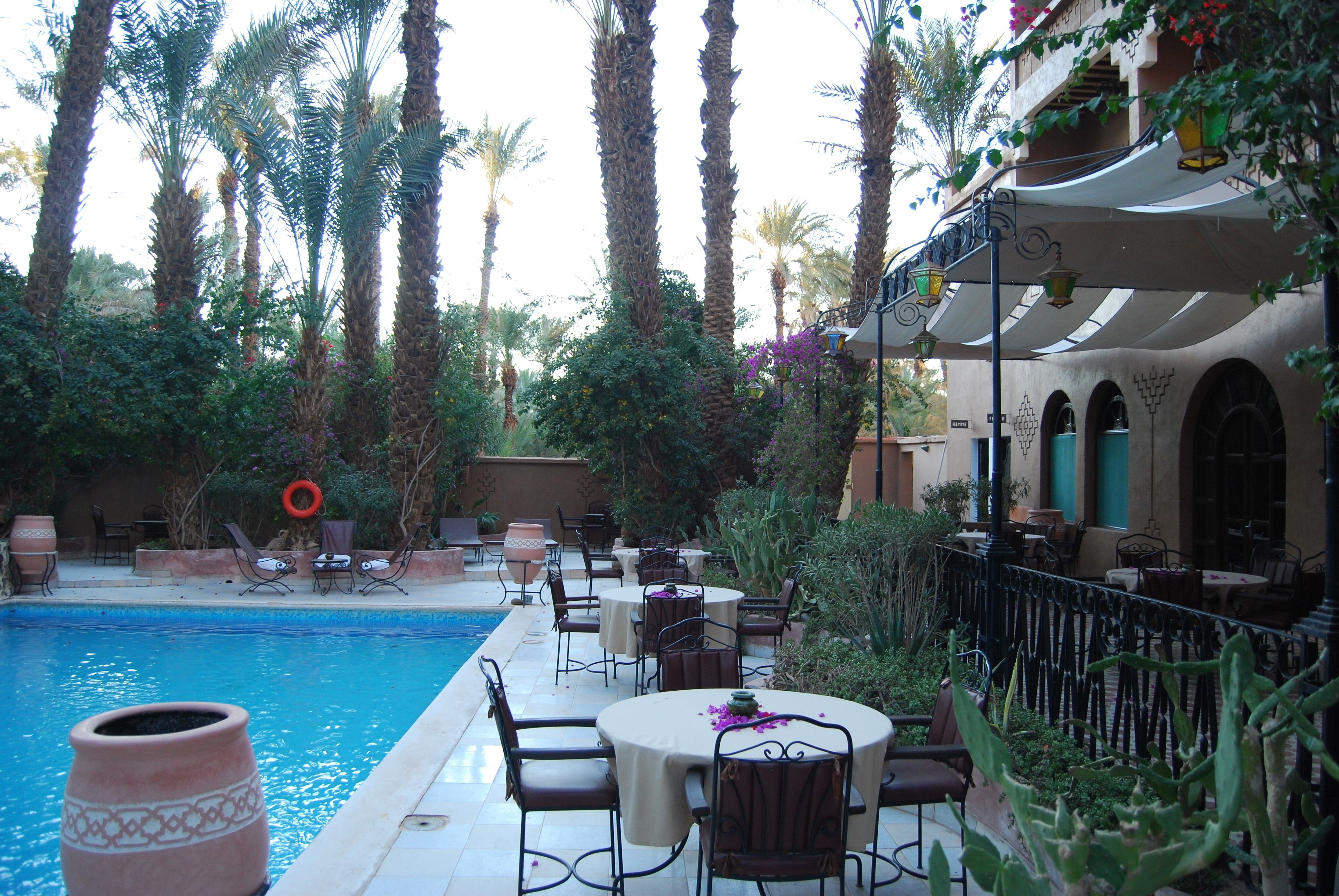 An outdoor pool area with palm trees, tables, and chairs, surrounded by lush greenery and a building with arches.