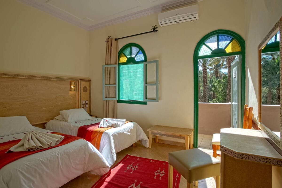 Habitación de hotel con dos camas, un pequeño banco, un escritorio y un balcón con vistas.