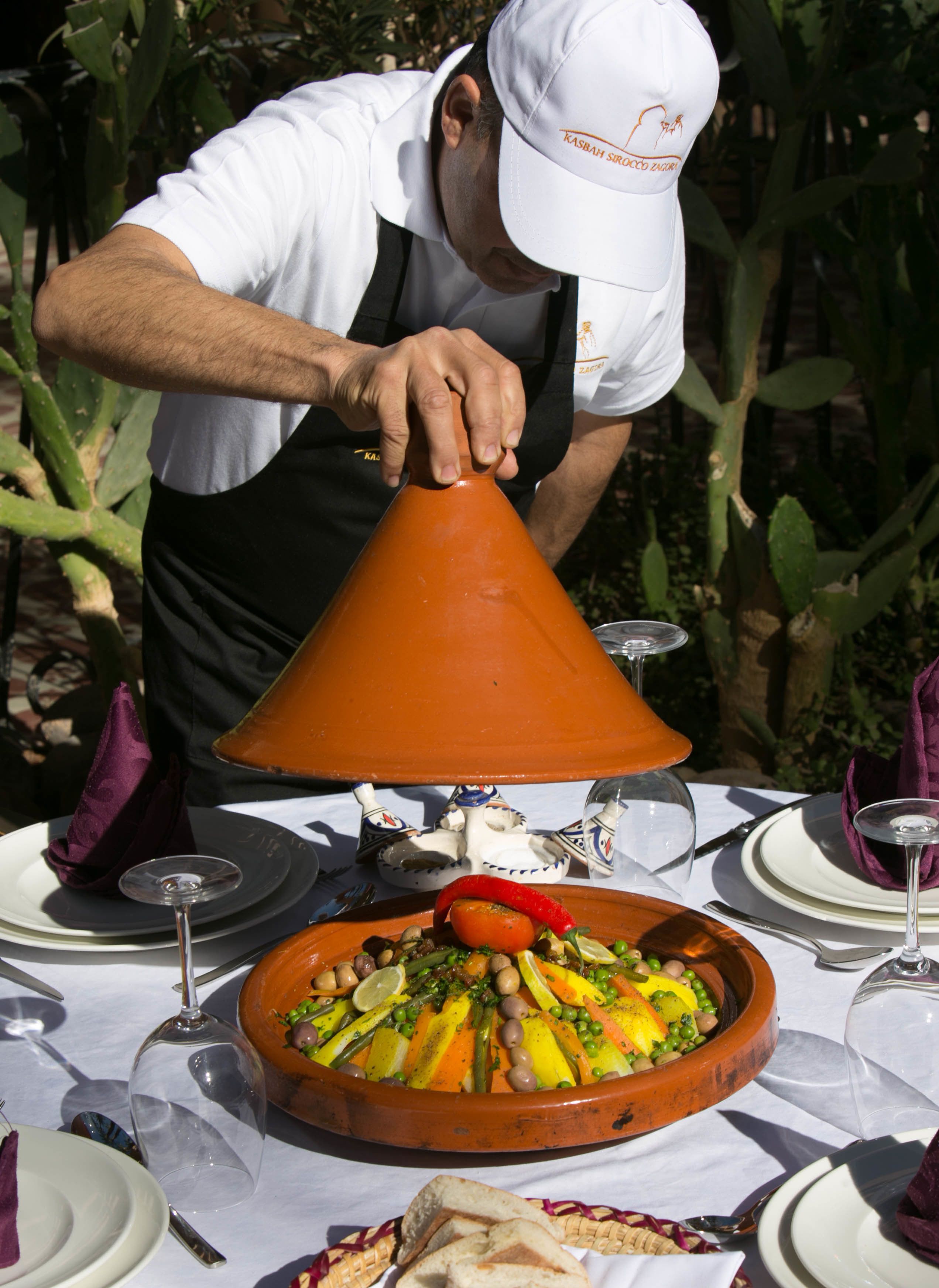 A person serving food from a traditional clay pot at an outdoor dining table with plates, glasses, and a napkin.