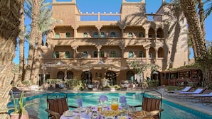 A poolside dining area with a table set for breakfast, overlooking a pool and a large building with multiple balconies. - Kasbah Sirocco (Zagora)