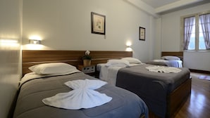 Habitación de hotel con dos camas, cada una con una almohada y una toalla. Hay una mesita de noche con una lámpara y una planta pequeña. La habitación tiene un cabecero de madera y una ventana con cortinas.