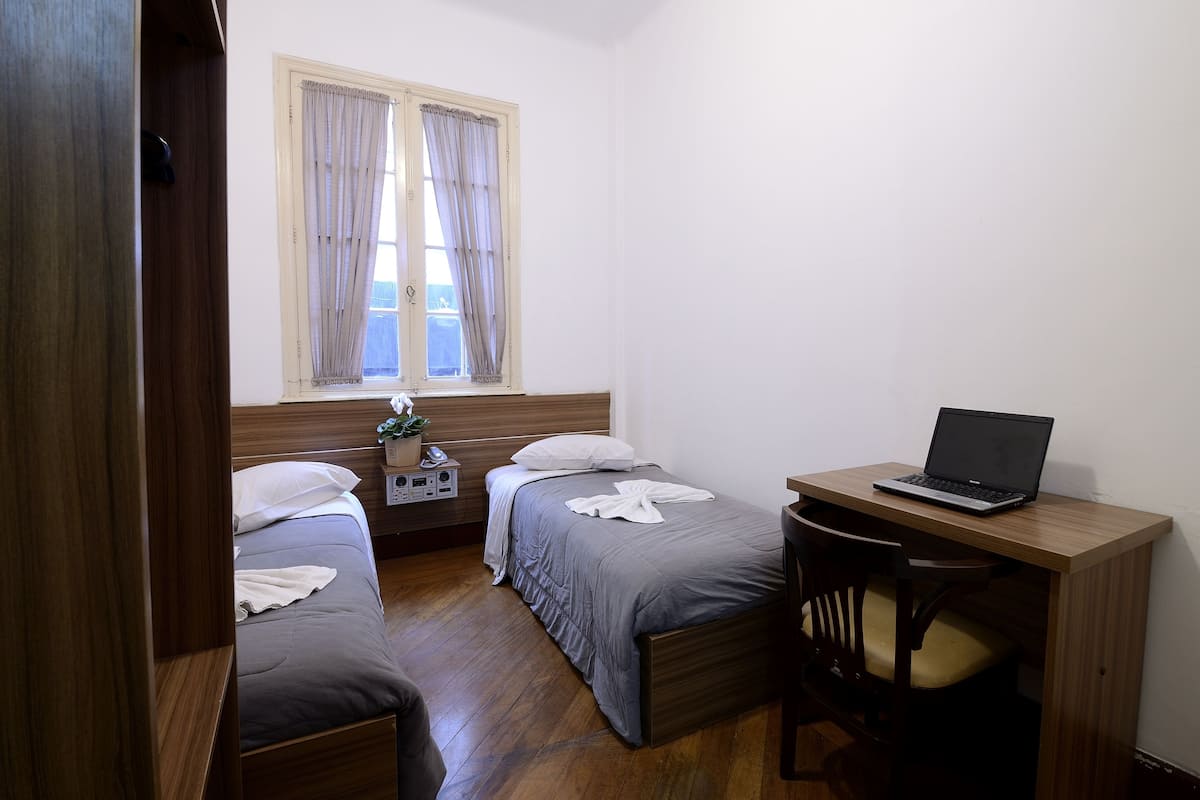 Ein Schlafzimmer mit zwei Betten, einem Schreibtisch mit Laptop, einem Nachttisch mit Pflanze und einem Fenster mit Vorhängen.