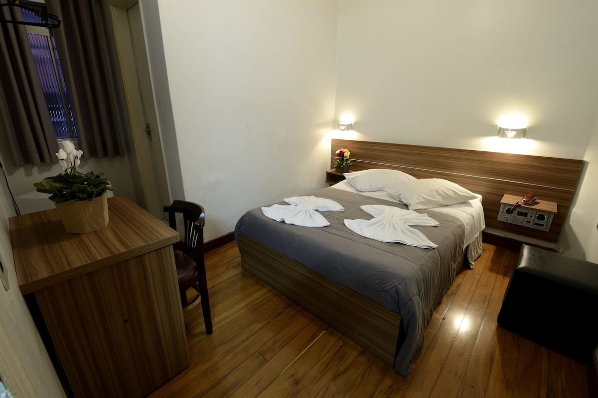 Ein Hotelzimmer mit hölzernem Kopfteil, einem Bett mit weißen Handtüchern, einem Nachttisch mit Lampe, einem Holztisch und einer Topfpflanze.