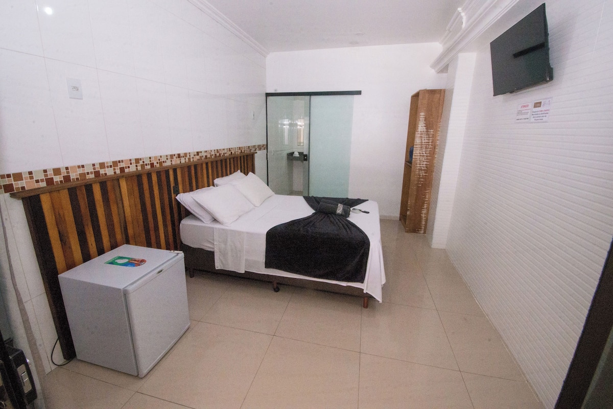 A small, clean hotel room with a bed, a small refrigerator, a TV mounted on the wall, and a sliding glass door leading to a bathroom.