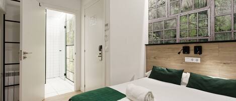 A hotel room with a bed, a green bedspread, a window with a view of greenery, a wooden headboard, and a door leading to a bathroom.