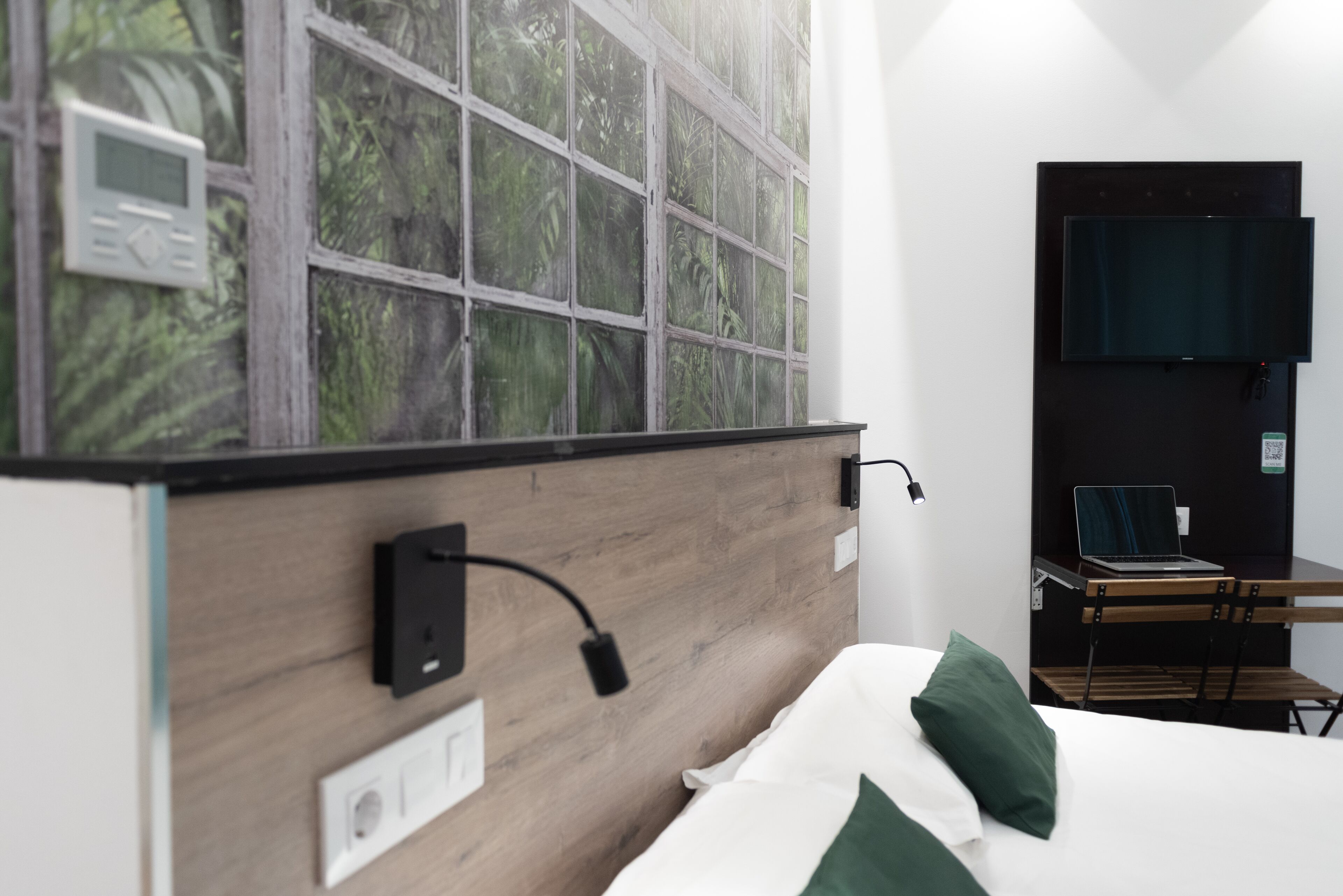 Habitación de hotel moderna con cabecera de madera, un ventanal con vistas a zonas verdes, televisor de pantalla plana en la pared y un escritorio con una computadora portátil.