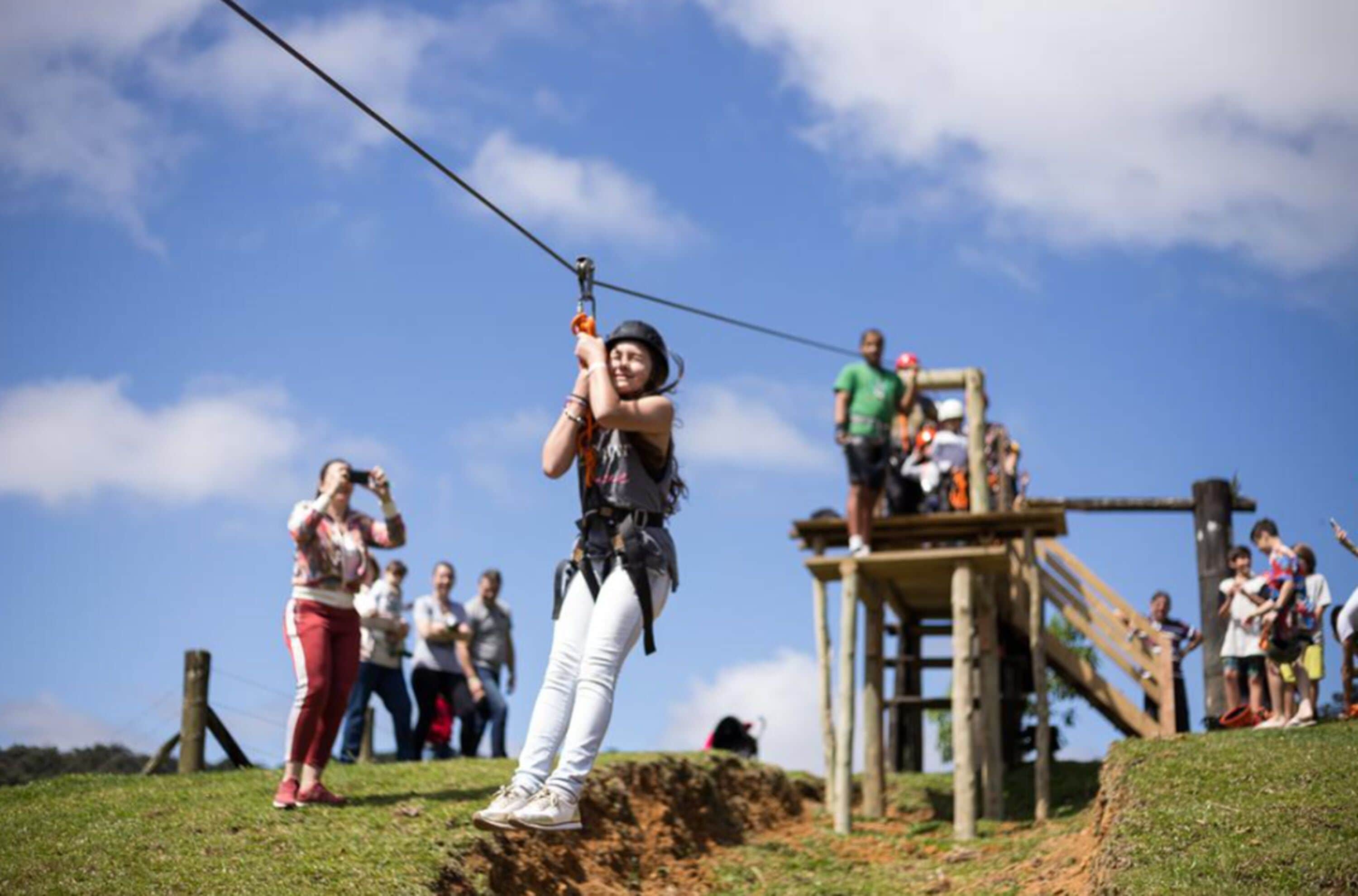 Uma pessoa fazendo tirolesa presa por um arnês de segurança, com uma plataforma de madeira e céu claro.
