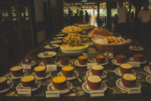 Une table de buffet proposant divers plats, chacun étant accompagné d’une étiquette indiquant son nom, notamment « Margarina », « Manteiga », « Requeijão » et « Nata ».