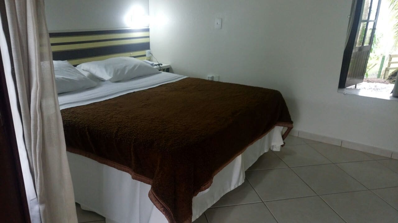 Una cama con una manta marrón y sábanas blancas, una mesita de noche con una lámpara y una ventana con cortinas.