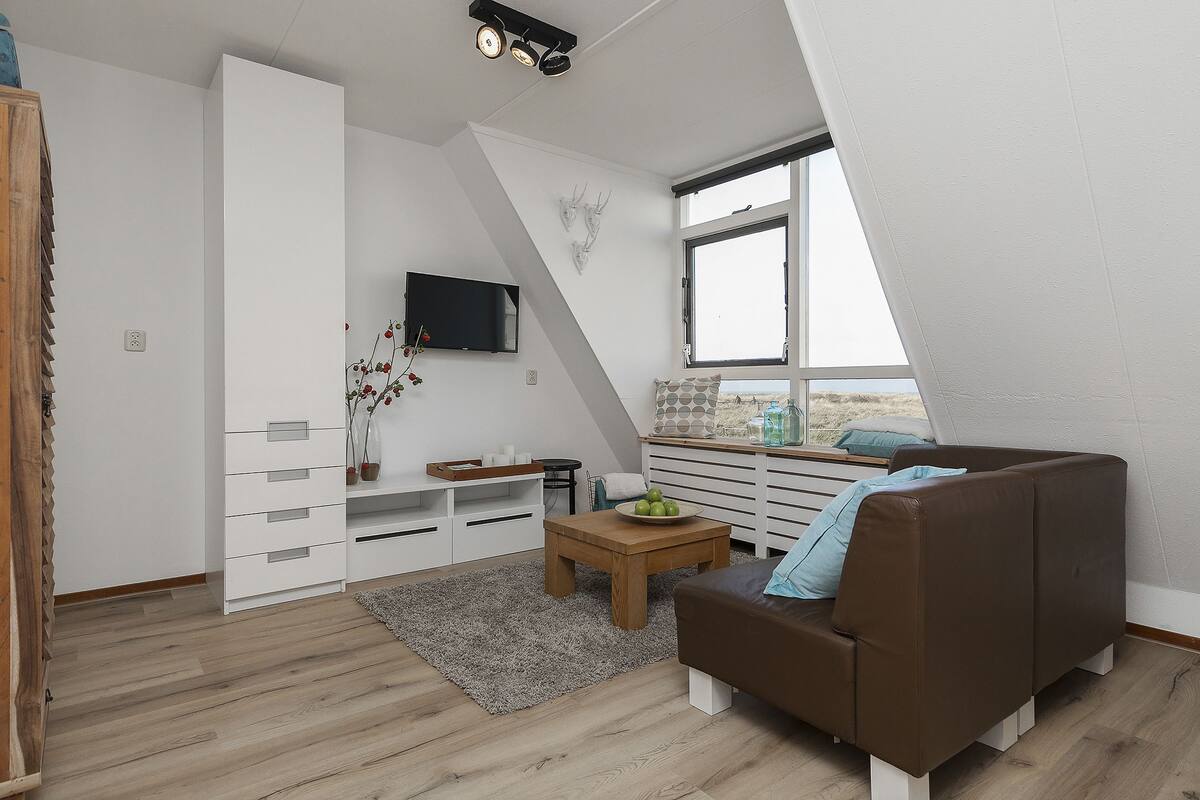 Ein modernes Schlafzimmer im Loft-Stil mit schrägen Wänden, einem braunen Ledersofa, einem Holztisch und einem großen Fenster mit Aussicht.
