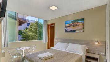 A bedroom with a bed, a nightstand, a wall-mounted lamp, a window with a view of greenery, and a painting on the wall.