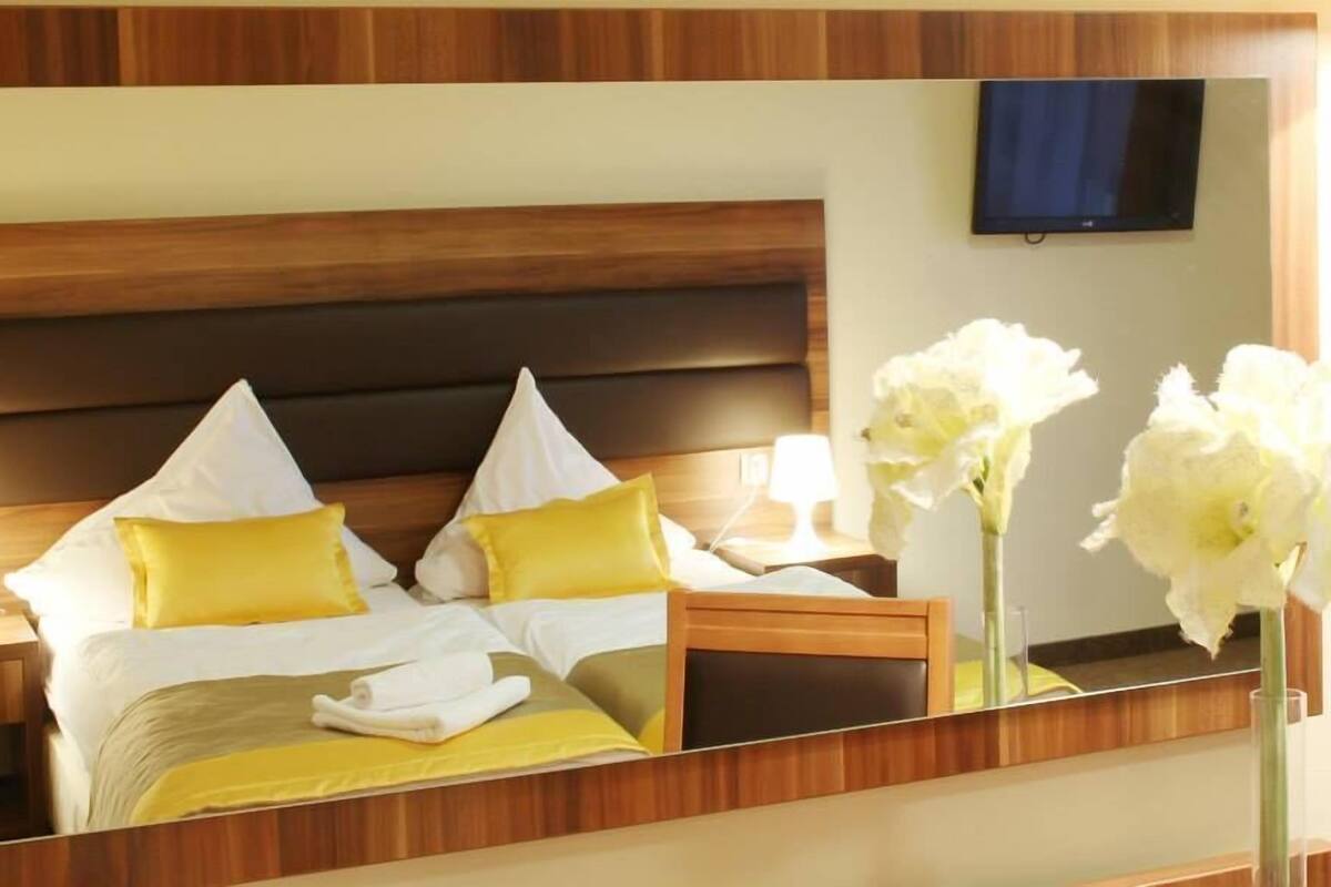 Une chambre d’hôtel avec une tête de lit en bois, deux lits avec une literie jaune et blanche, une table de chevet avec une lampe et un vase contenant des fleurs blanches.