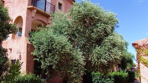 Ein Gebäude mit rosafarbenem Mauerwerk, einem Balkon und einem gepflegten Garten mit verschiedenen Bäumen und Sträuchern.