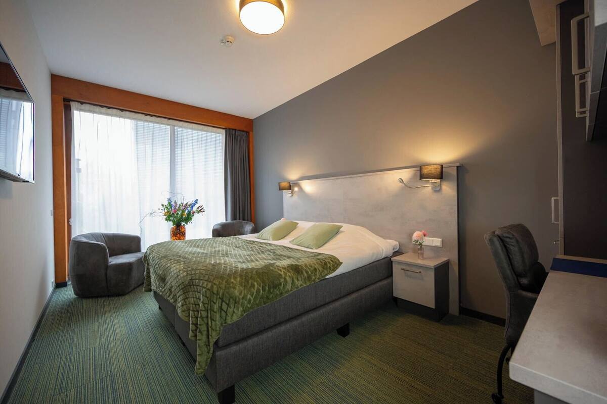 A hotel room with a bed, a desk, a chair, a nightstand, a wall-mounted lamp, and a window with curtains.