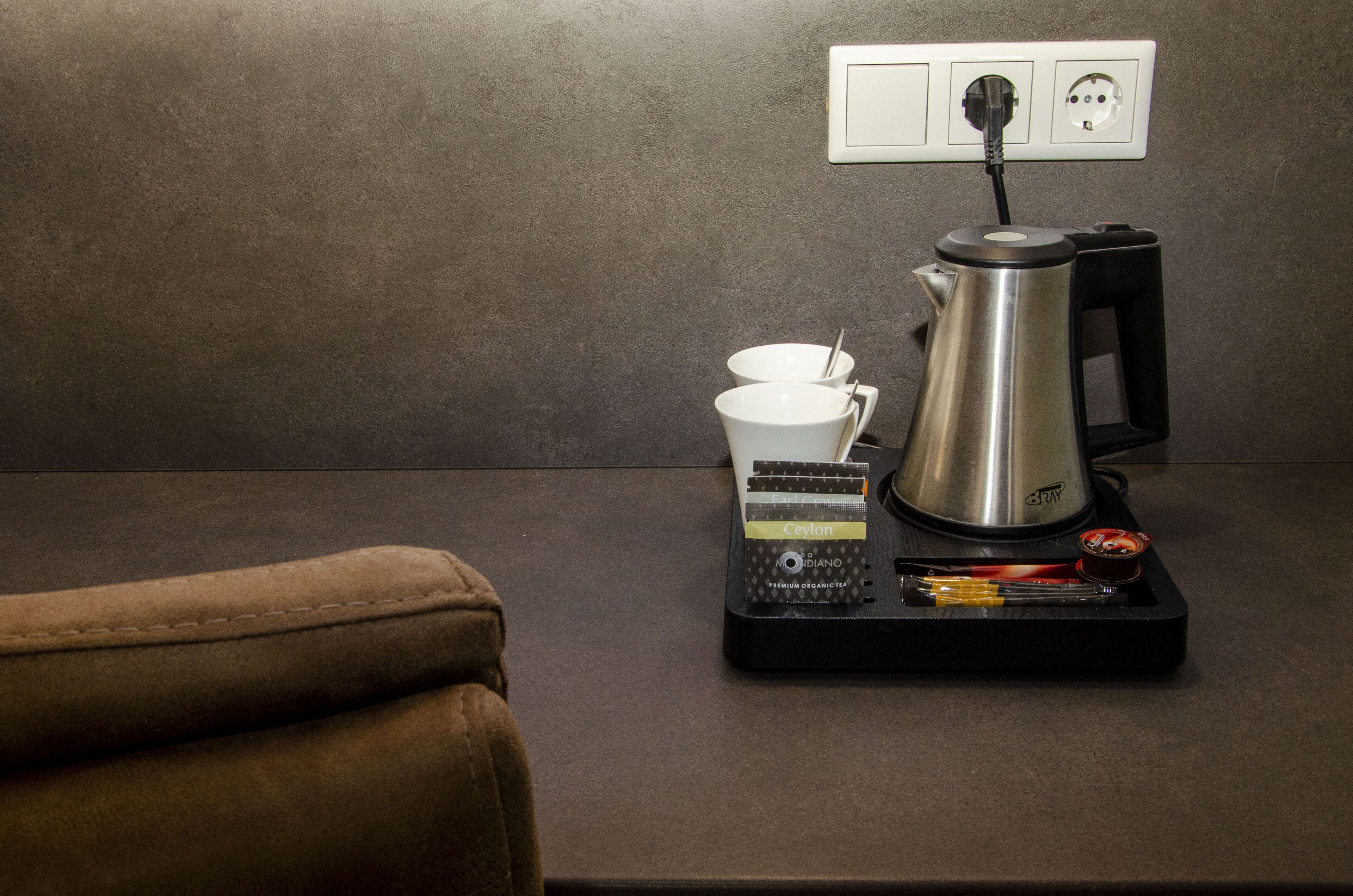 A coffee maker on a table with a power outlet, a cup, and a spoon.