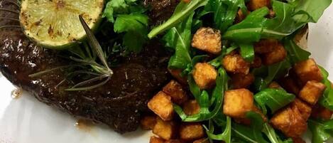 Une assiette comprenant un steak grillé, une tranche de citron, de la roquette et des croûtons grillés.