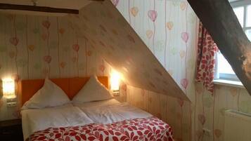 Un dormitorio con techo inclinado, cama con ropa de cama blanca y roja, y papel tapiz estampado.