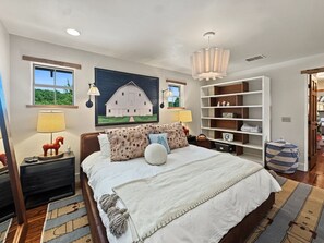 Un dormitorio con una cama grande, una mesita de noche, una lámpara, un estante en la pared y un cuadro de un granero.