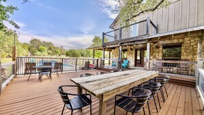 A wooden deck with a long table and chairs, a pool, and a stone building with a covered patio area.